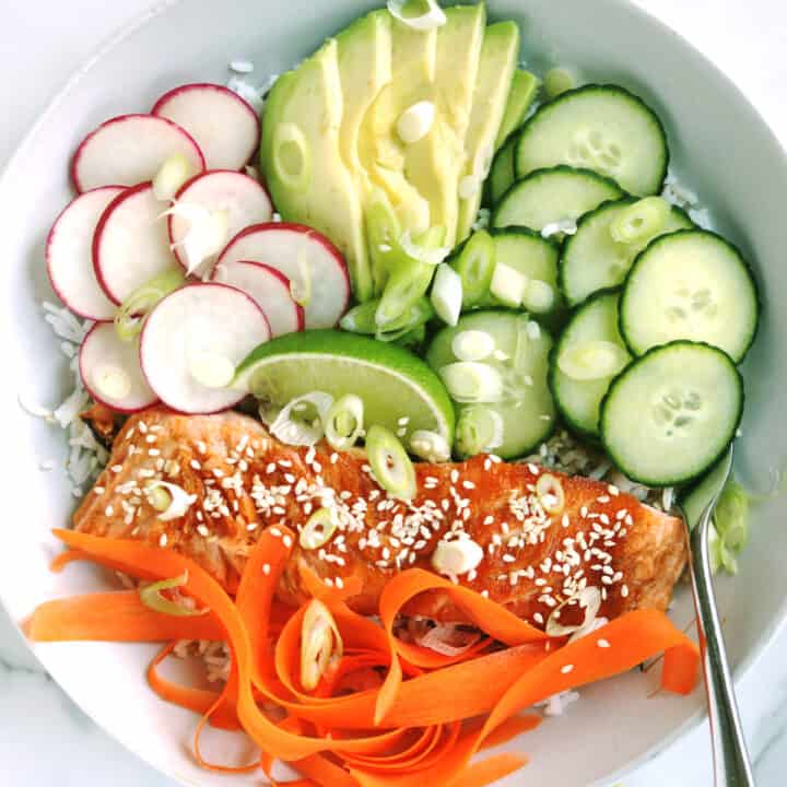 A salmon bowl with rice, seared salmon, sliced cucumber, avocado, radish, carrot ribbons, lime wedge, and sprinkled with sesame seeds and chopped green onions, with a fork on the side.