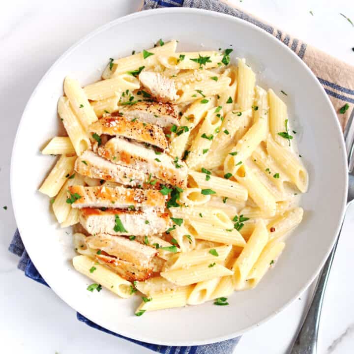 A white bowl filled with creamy penne pasta, topped with sliced grilled chicken breast and garnished with chopped parsley and grated cheese. A fork is beside the bowl on a marble countertop.
