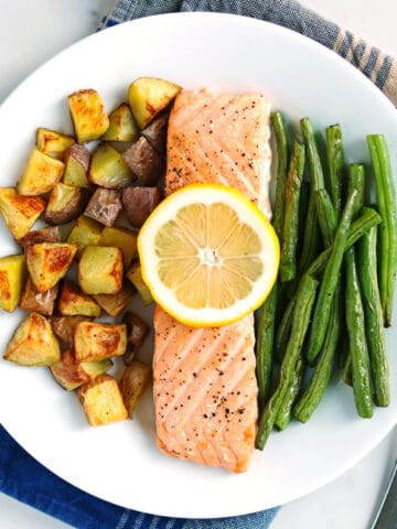 A white plate with baked salmon topped with a lemon slice and roasted potato and green beans, set on a blue striped napkin. A fork is placed beside the plate.