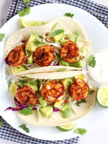 Two shrimp tacos topped with avocado, slaw, and cilantro are served on a white plate with lime wedges and a side of creamy sauce, placed on a blue and white checkered cloth.