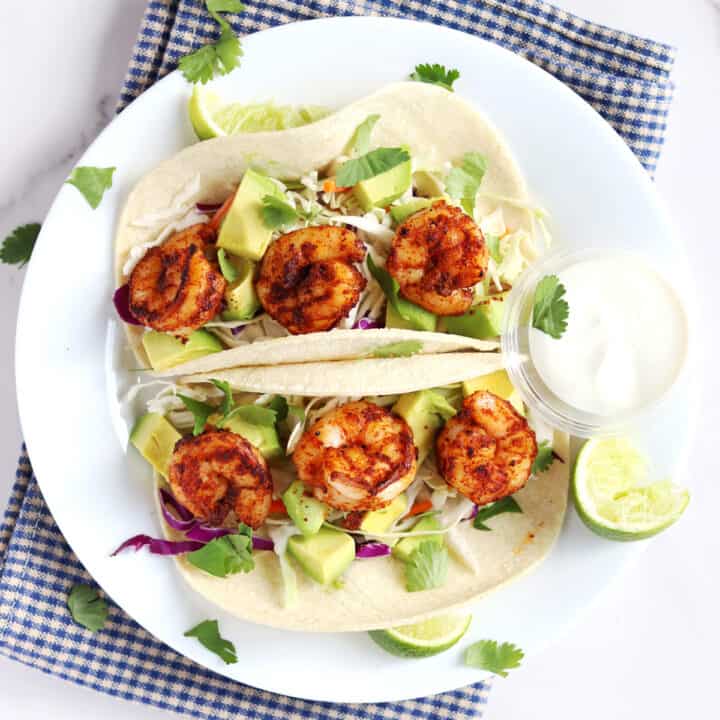 Two shrimp tacos topped with avocado, slaw, and cilantro are served on a white plate with lime wedges and a side of creamy sauce, placed on a blue and white checkered cloth.