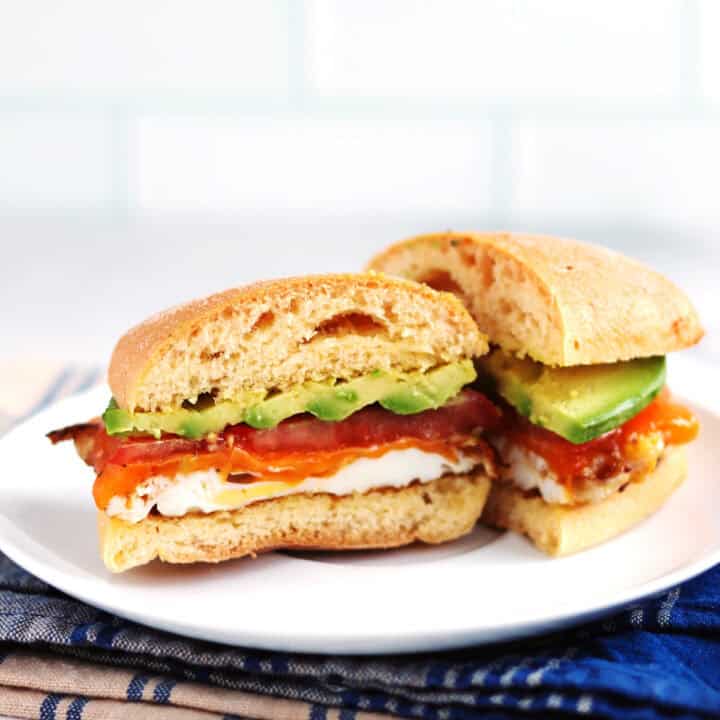 A breakfast sandwich on ciabatta cut in half, filled with egg, melted cheese, tomato, and avocado slices, placed on a white plate with a blue and beige napkin underneath.