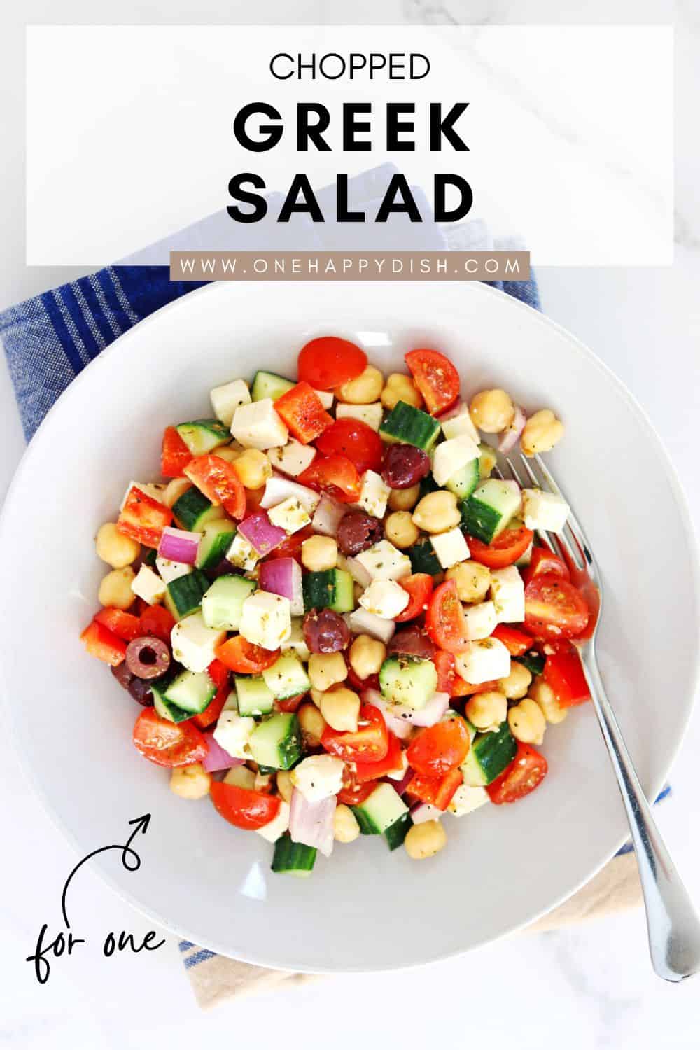 A white bowl filled with chopped Greek salad, including cucumbers, tomatoes, red onion, chickpeas, olives, and feta cheese, sits on a blue and tan napkin with a fork beside it. Text above reads “Chopped Greek Salad.”.