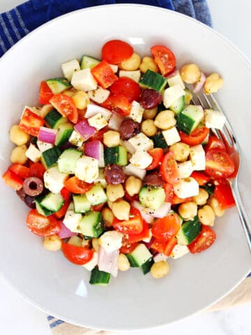 A bowl of colorful chopped Greek salad with cherry tomatoes, cucumber, red onion, red bell pepper, Kalamata olives, chickpeas, and feta cheese cubes, served with a fork on a blue-striped napkin.