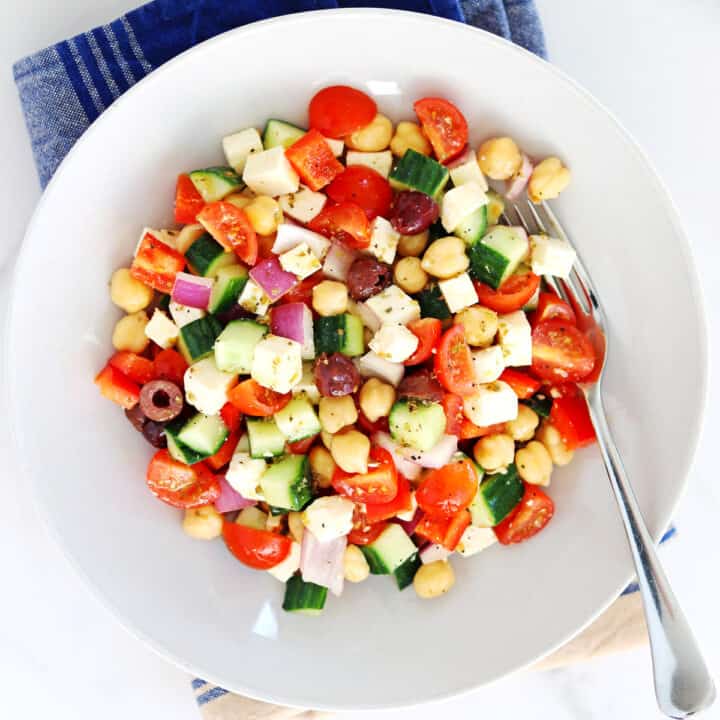 A bowl of colorful chopped Greek salad with cherry tomatoes, cucumber, red onion, red bell pepper, Kalamata olives, chickpeas, and feta cheese cubes, served with a fork on a blue-striped napkin.
