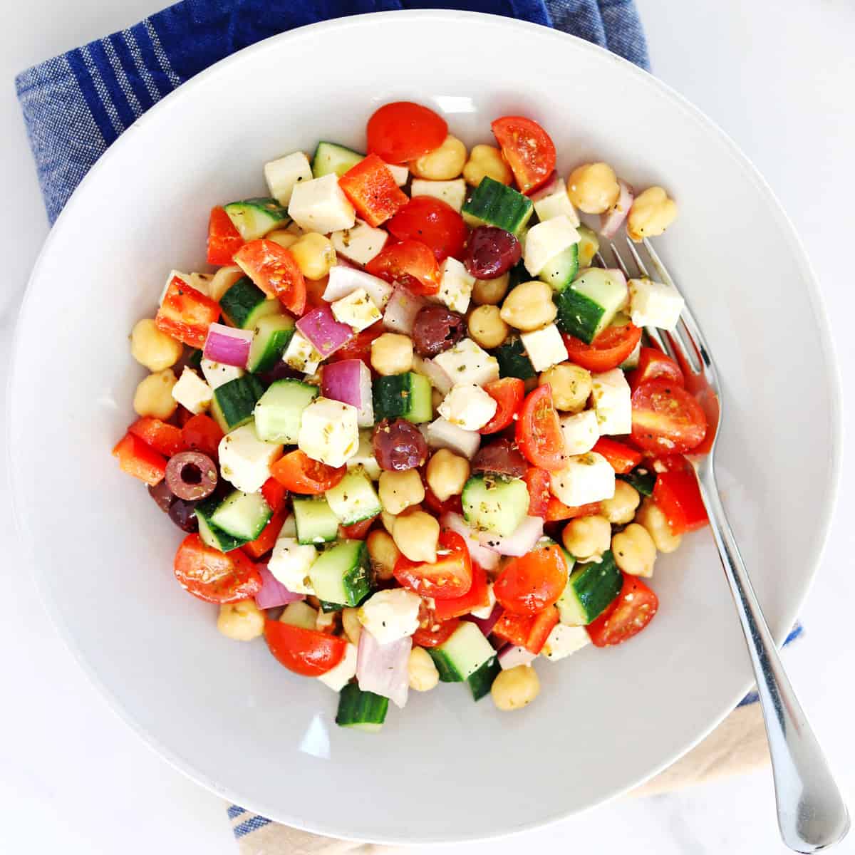 A bowl of colorful chopped Greek salad with cherry tomatoes, cucumber, red onion, red bell pepper, Kalamata olives, chickpeas, and feta cheese cubes, served with a fork on a blue-striped napkin.