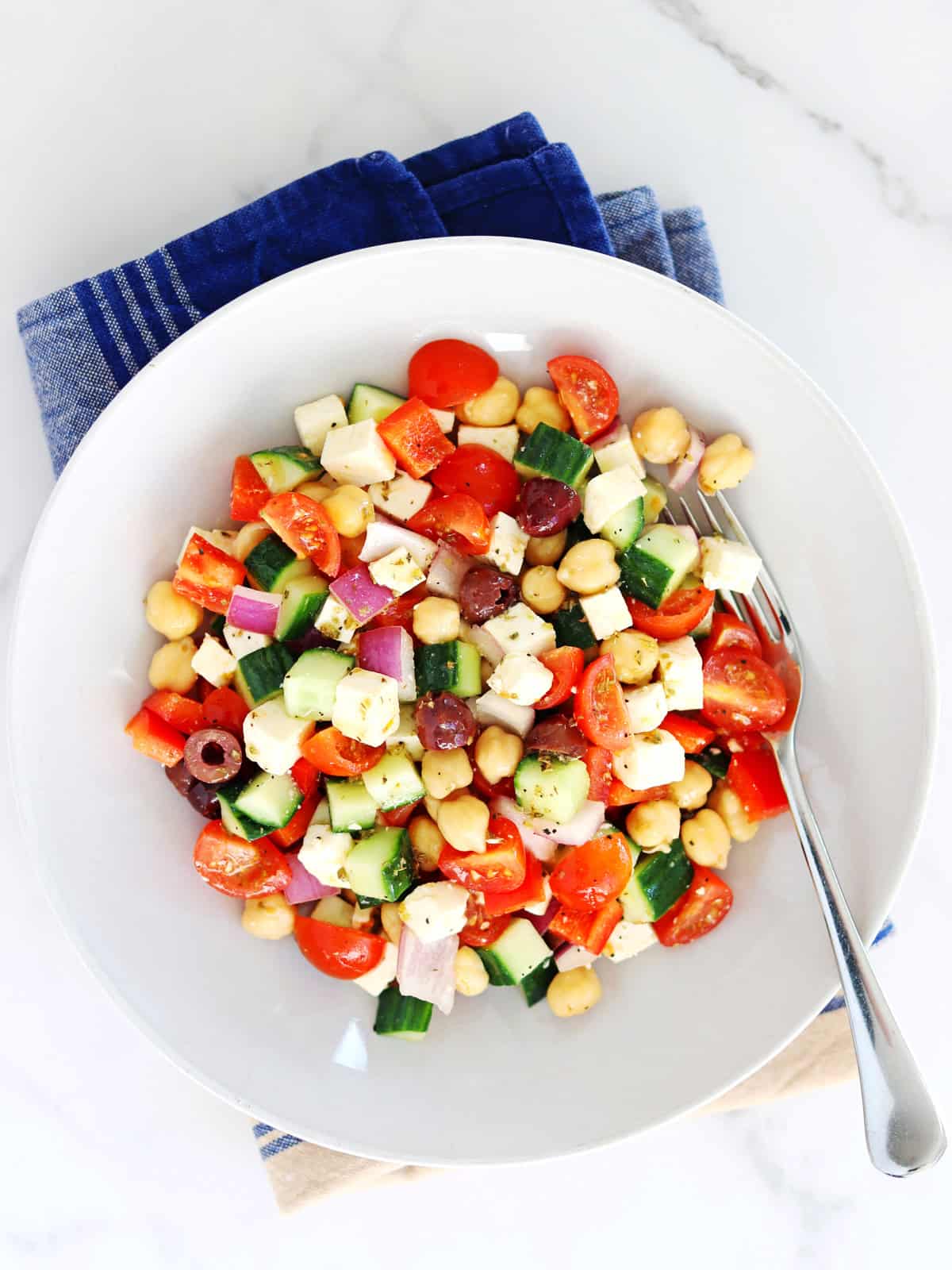 Chopped Greek salad with cherry tomatoes, cucumbers, chickpeas, red onion, feta cheese, and olives sits on a blue and tan napkin, with a fork resting on the side.