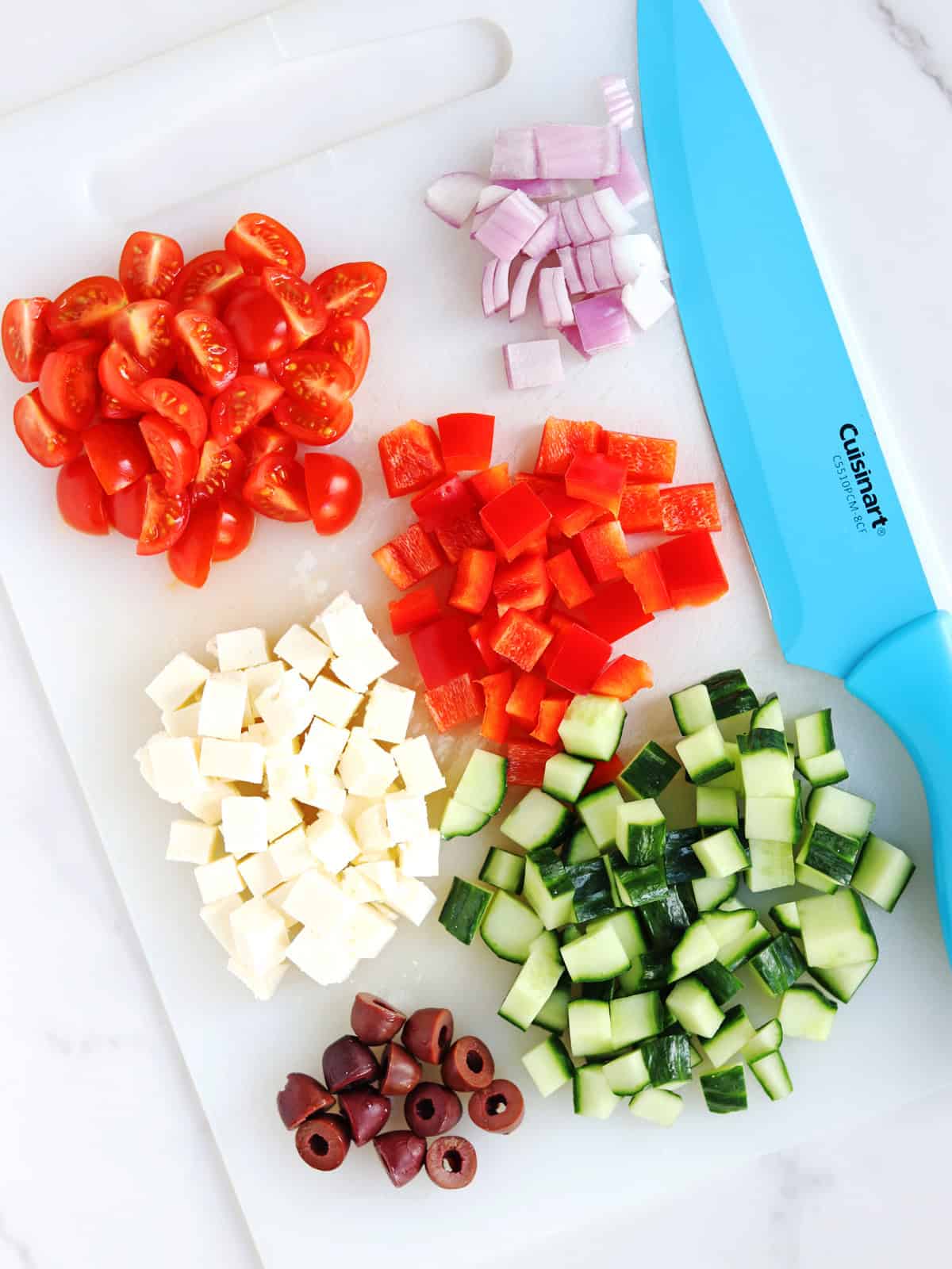 A cutting board with piles of chopped cherry tomatoes, red onion, red bell pepper, cucumber, feta cheese, and Kalamata olives, next to a blue kitchen knife.