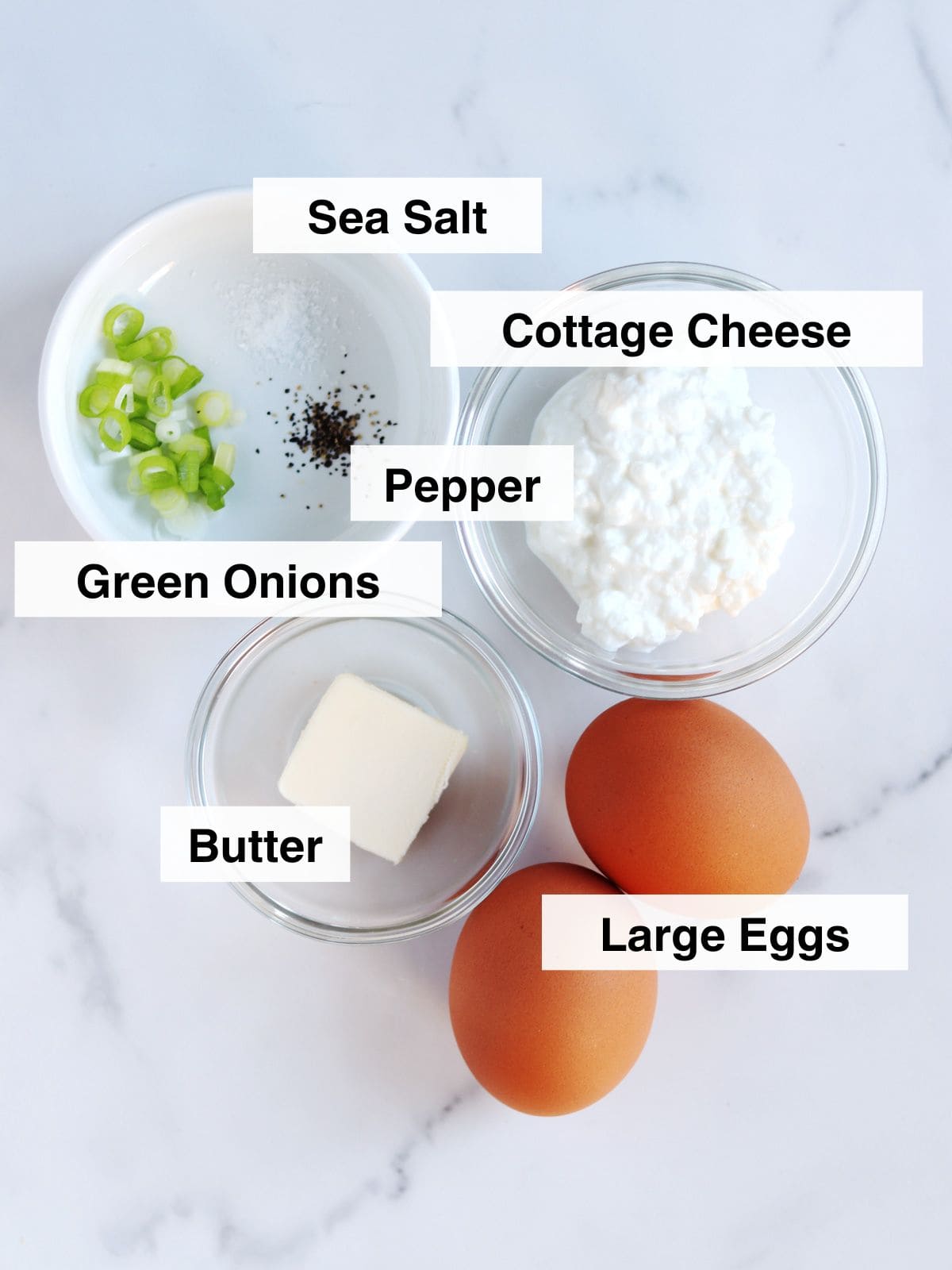 Ingredients for a recipe arranged on a white surface: a bowl of cottage cheese, two large eggs, a dish with chopped green onions, butter in a small bowl, and a pinch of sea salt and pepper, each labeled.