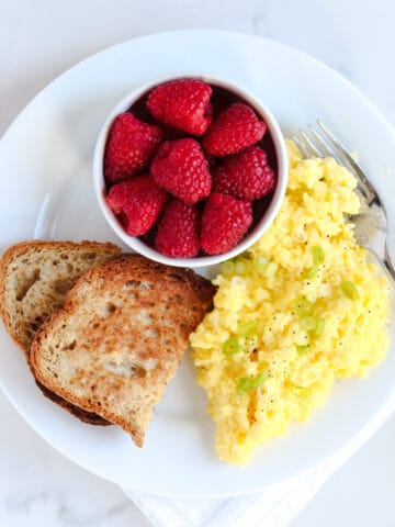 A white plate with cottage cheese scrambled eggs topped with chopped green onions, two slices of toasted bread, and a small bowl of fresh raspberries, with a fork on the side.