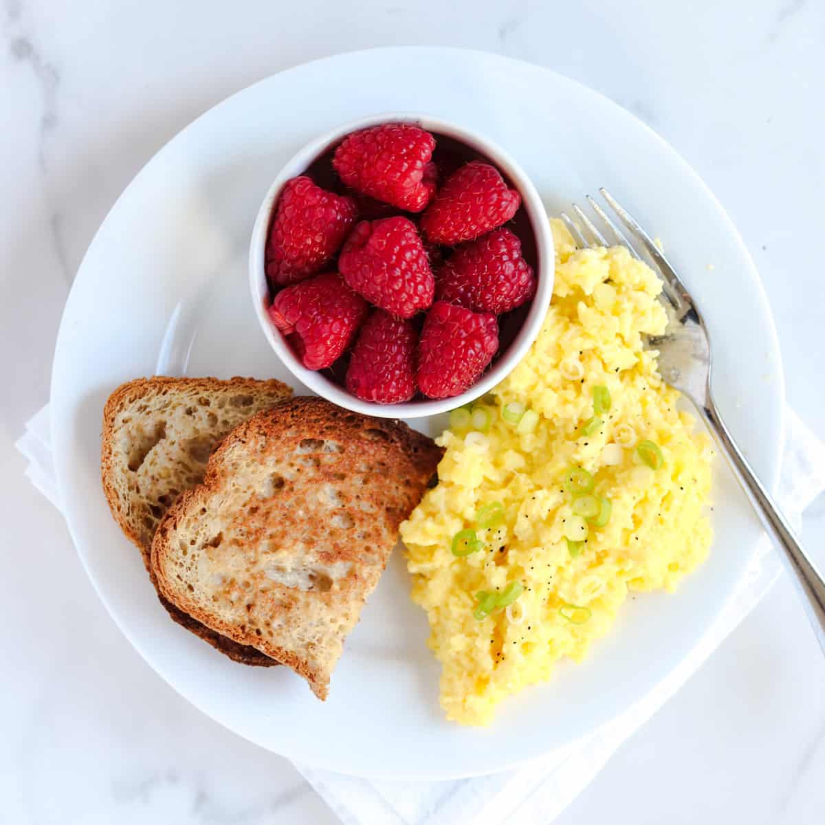 A white plate with cottage cheese scrambled eggs topped with chopped green onions, two slices of toasted bread, and a small bowl of fresh raspberries, with a fork on the side.