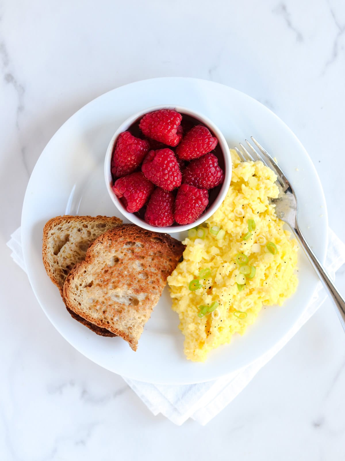 A white plate with cottage cheese scrambled eggs topped with green onions, two slices of toasted bread, and a small bowl of fresh raspberries, with a fork on the side, on a white marble surface.