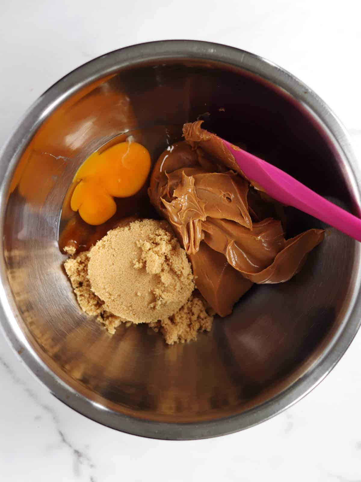 A metal mixing bowl containing an egg yolk, brown sugar, and peanut butter with a purple spatula resting on the side, set on a white marble countertop.