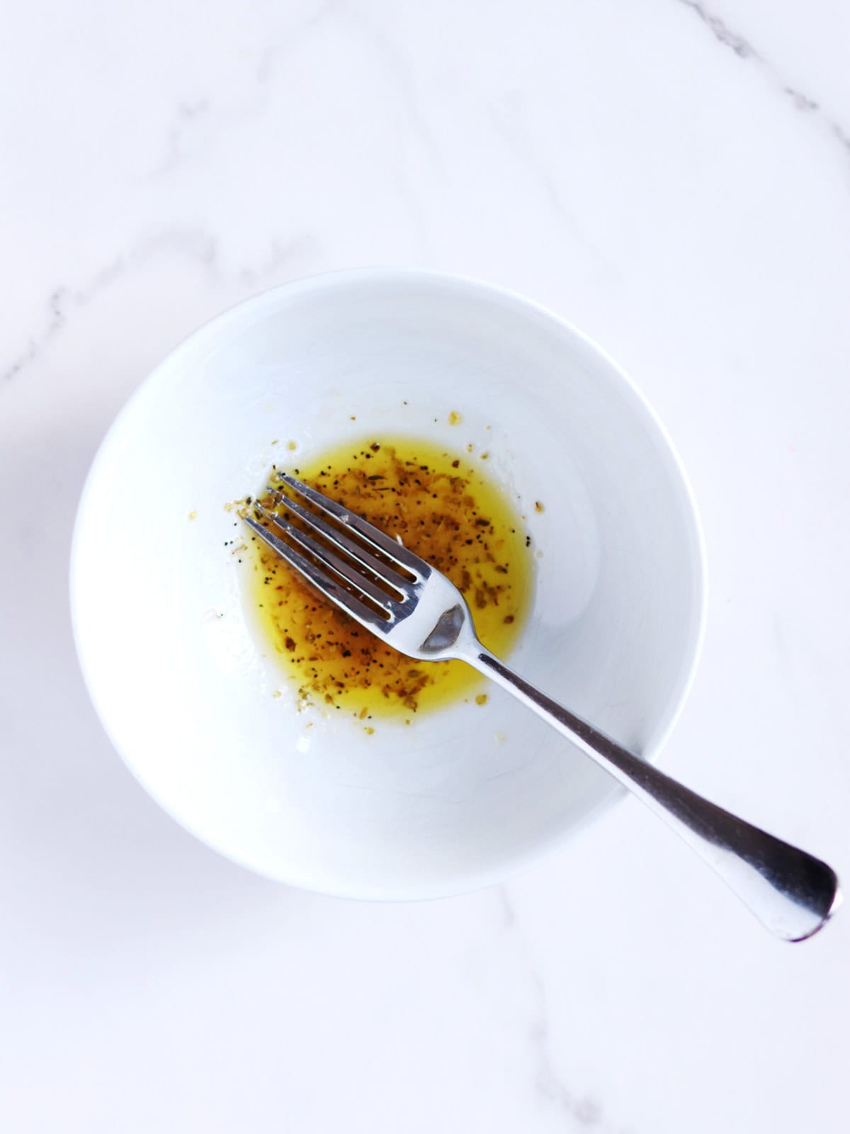 A white bowl containing a small amount of homemade Greek salad dressing, with a fork resting inside, sits on a white marble surface.