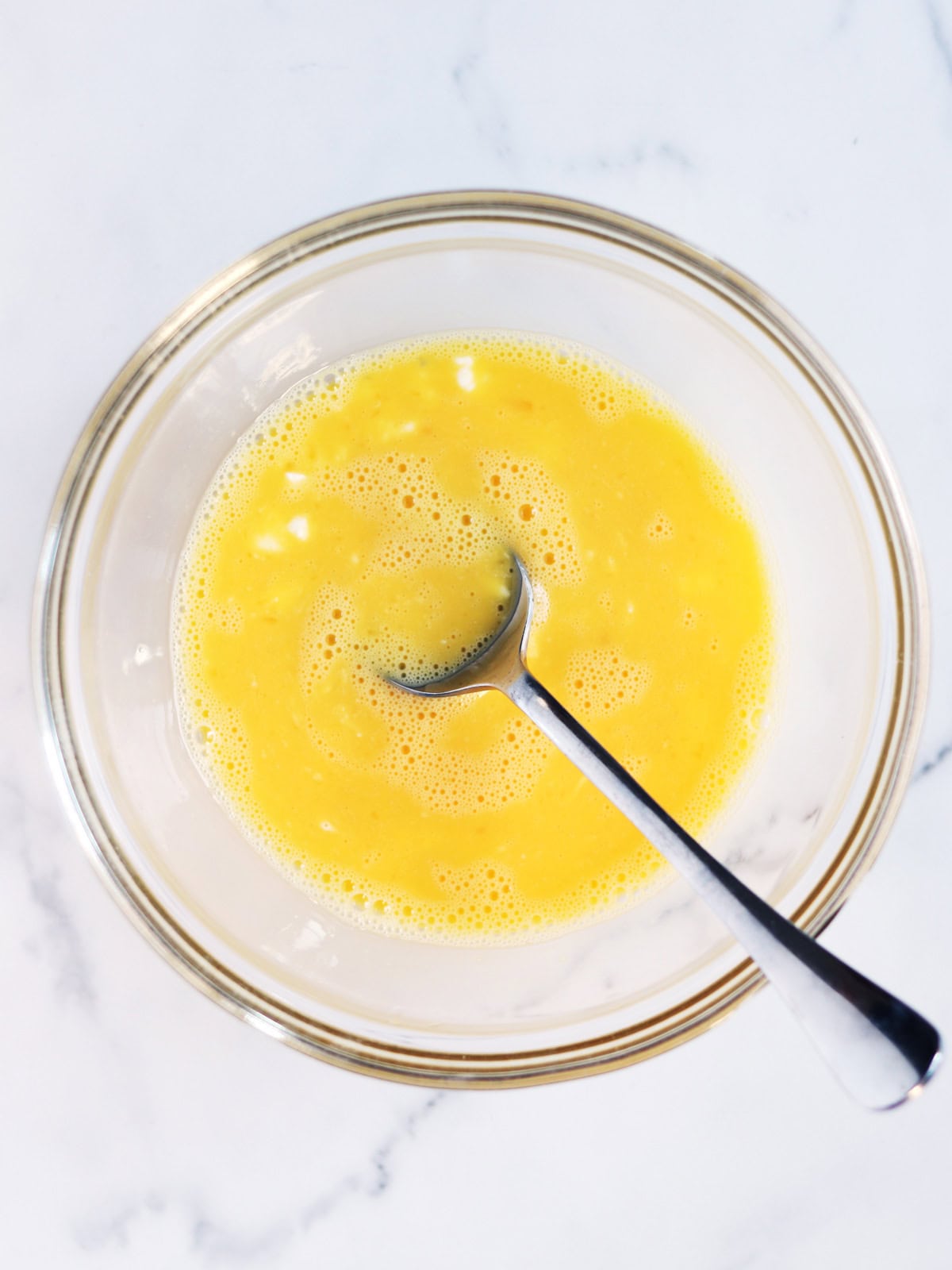 A glass bowl filled with beaten eggs, cottage cheese and a metal spoon resting inside, set on a white marble surface.