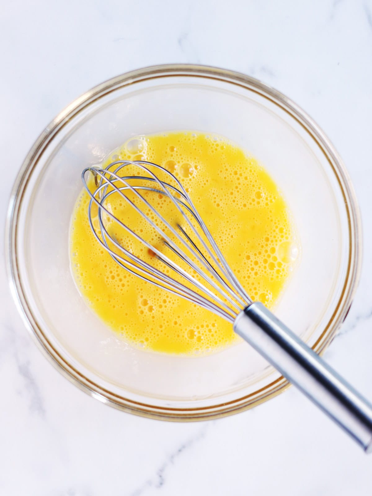 A metal whisk rests in a glass bowl filled with beaten eggs on a white marble surface.
