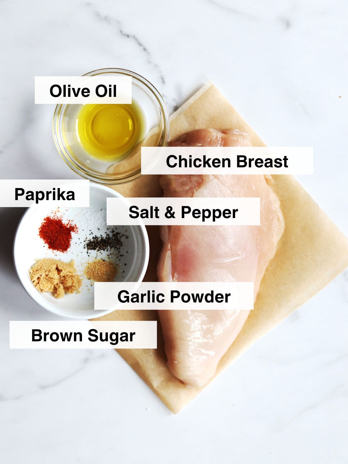 Ingredients for air fryer chicken breast including raw chicken breast on parchment paper beside small bowls of olive oil and mixed spices, labeled as olive oil, paprika, salt & pepper, brown sugar, and garlic powder, on a white marble countertop.