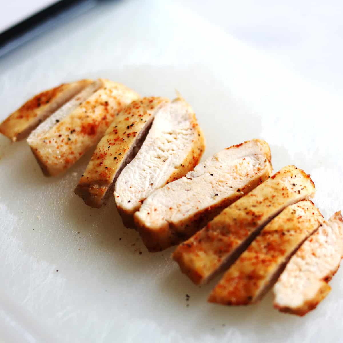 Sliced, cooked air fryer chicken breast seasoned with spices is arranged on a white cutting board. The chicken looks juicy and tender with a light golden-brown exterior.