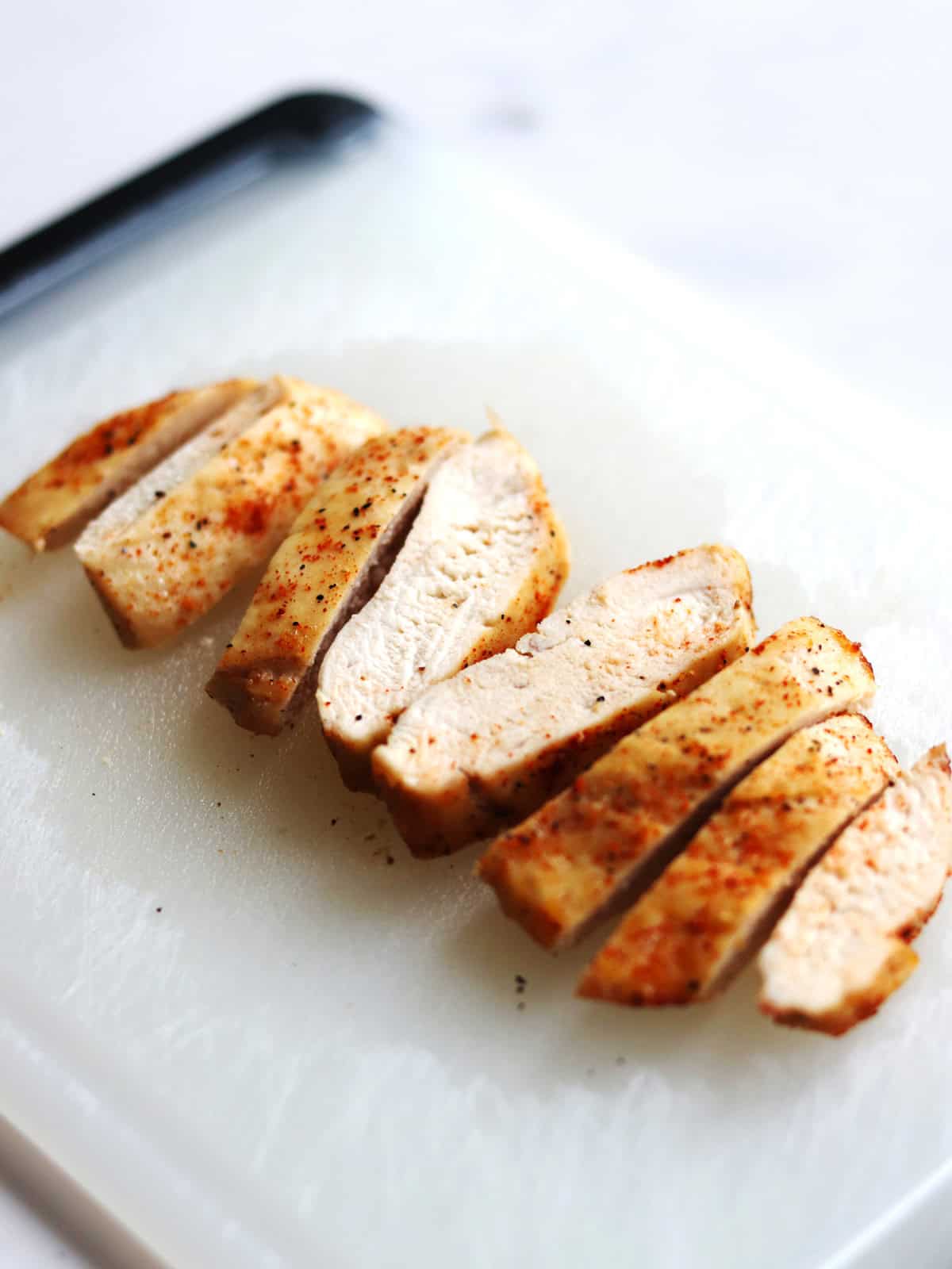 Sliced, cooked air fryer chicken breast seasoned with spices is arranged on a white cutting board.