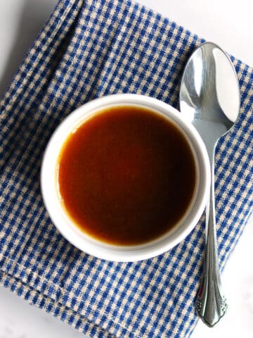A small white bowl filled with balsamic vinaigrette sits on a blue and white checkered napkin, with a silver spoon placed beside it on a white marble countertop.