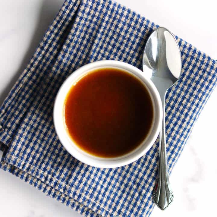 A small white bowl filled with balsamic vinaigrette sits on a blue and white checkered napkin, with a silver spoon placed beside it on a white marble countertop.