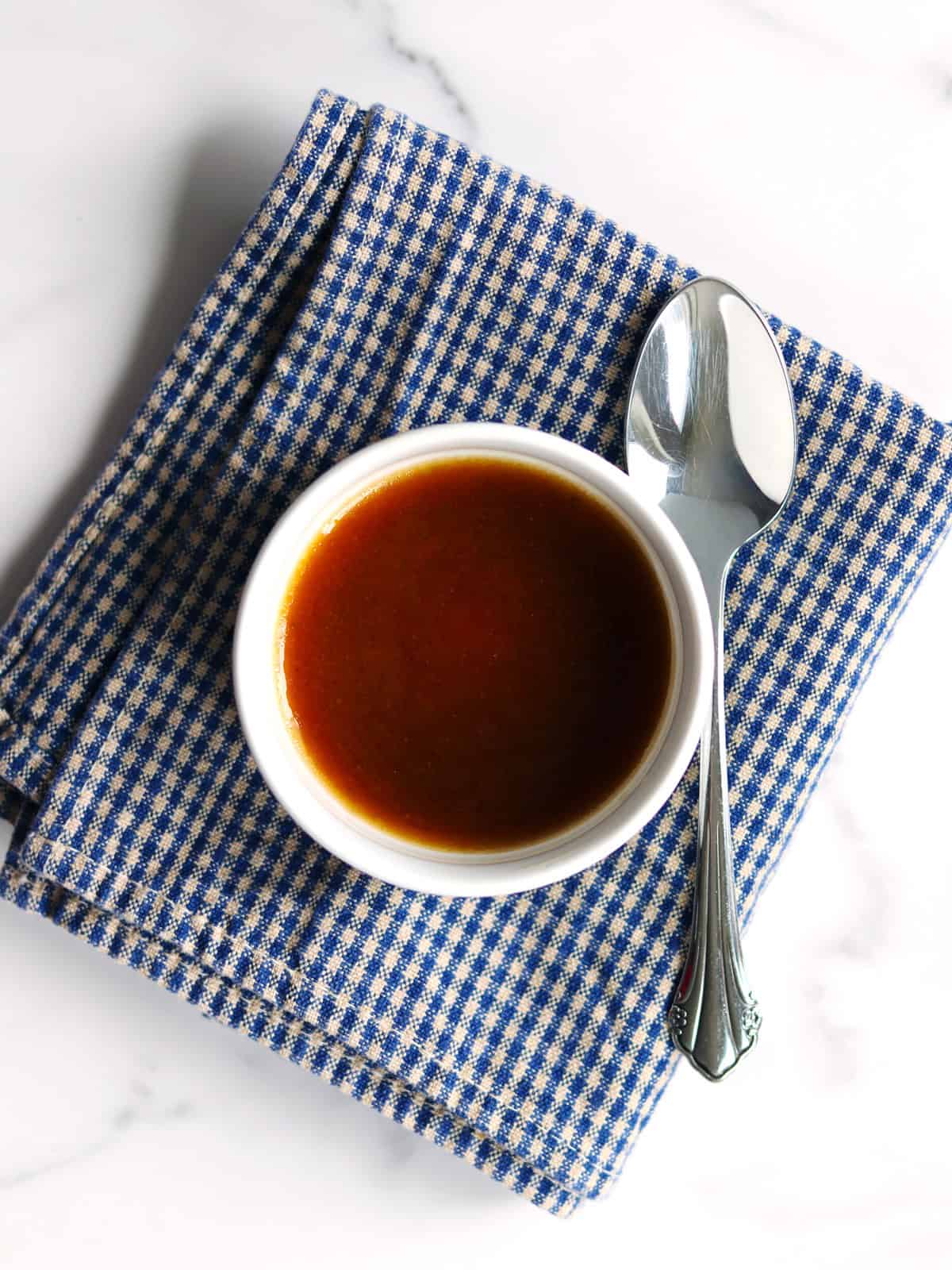 A small white bowl filled with balsamic vinaigrette is placed on a folded blue and white checkered napkin, with a silver spoon resting beside the bowl on a white marble countertop.