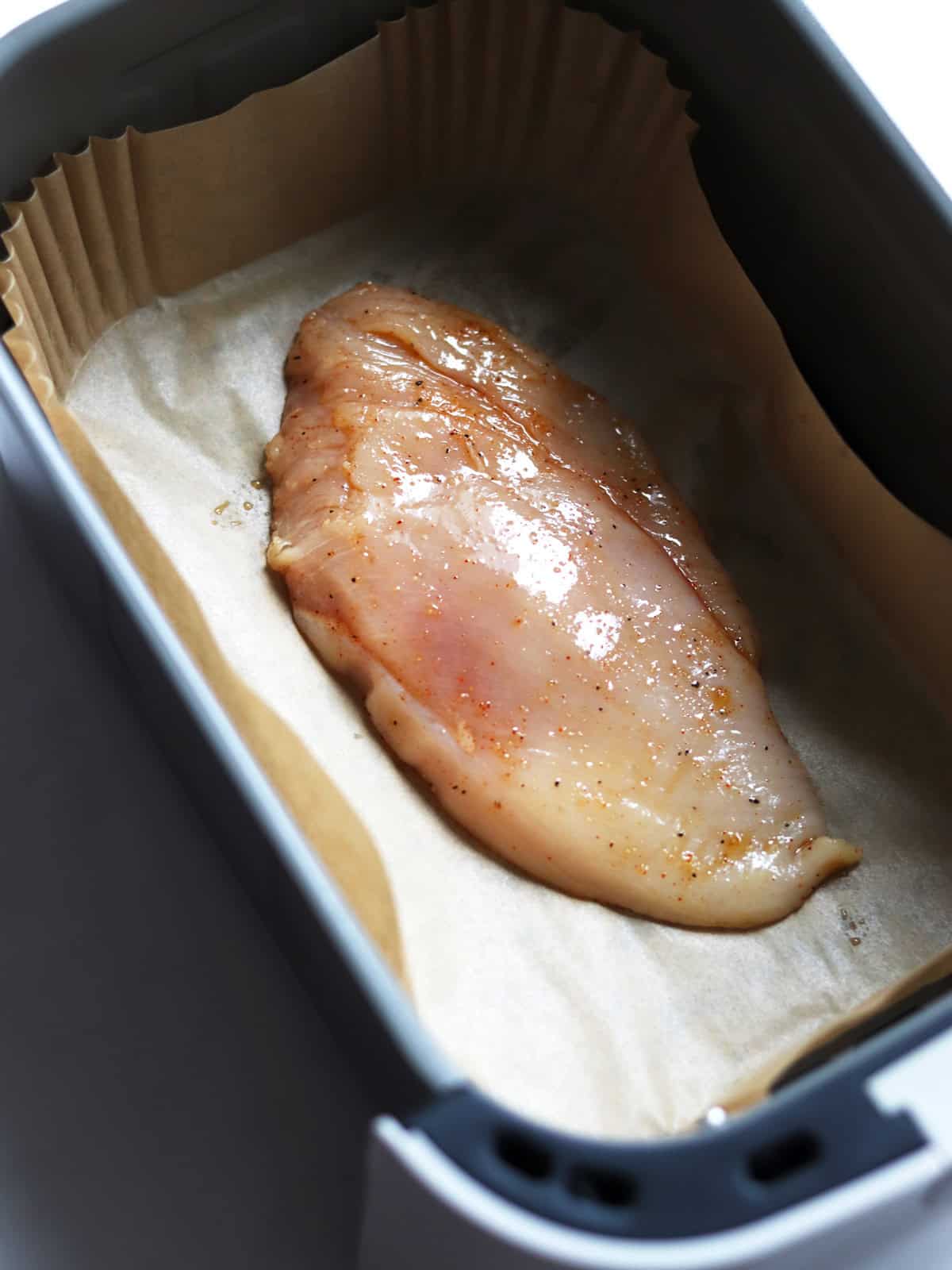 Raw, seasoned chicken breast placed on parchment paper inside an air fryer basket, ready for cooking.
