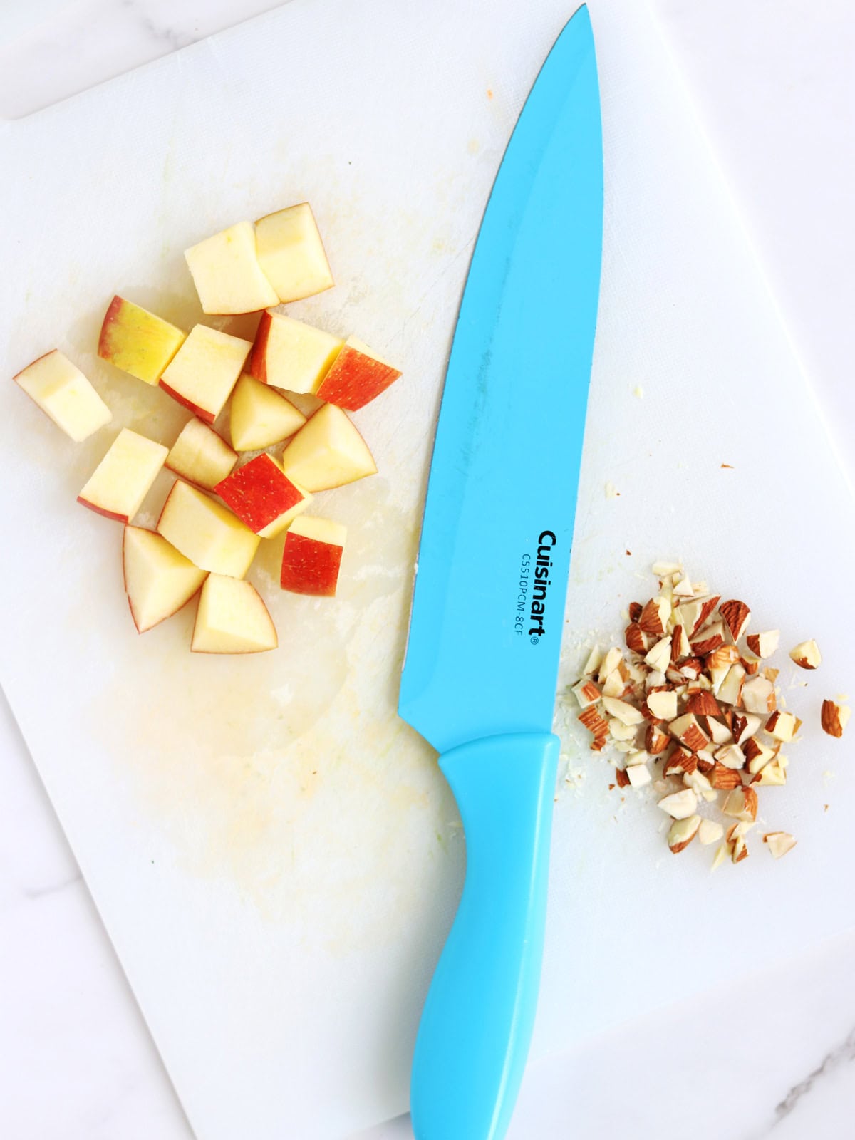A blue kitchen knife on a white cutting board with chopped red apple pieces on the left and chopped almonds on the right.