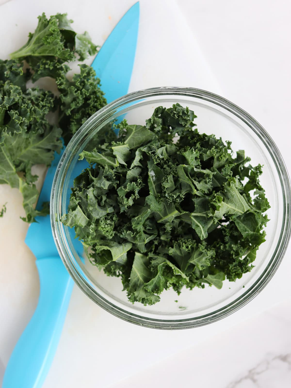 A clear glass bowl filled with chopped kale sits on a white cutting board next to a blue knife and more kale leaves.