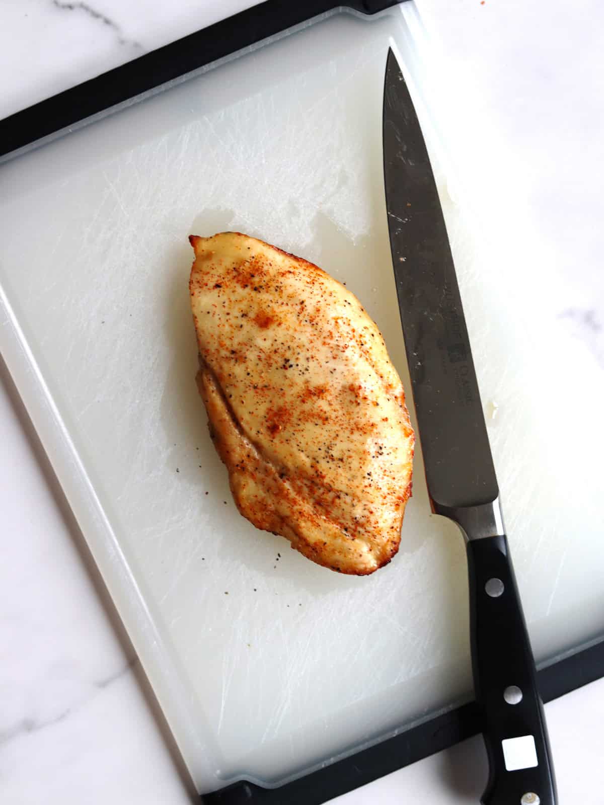 A seasoned cooked air fryer chicken breast sits on a white cutting board with a black-edged chef's knife beside it. The surface is lightly scratched, and the background is a white marble countertop.