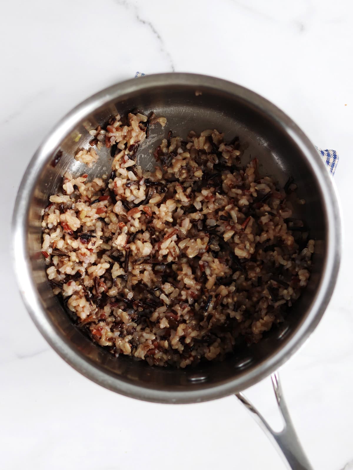 A stainless steel pot on a white marble surface contains a mixture of cooked wild rice.