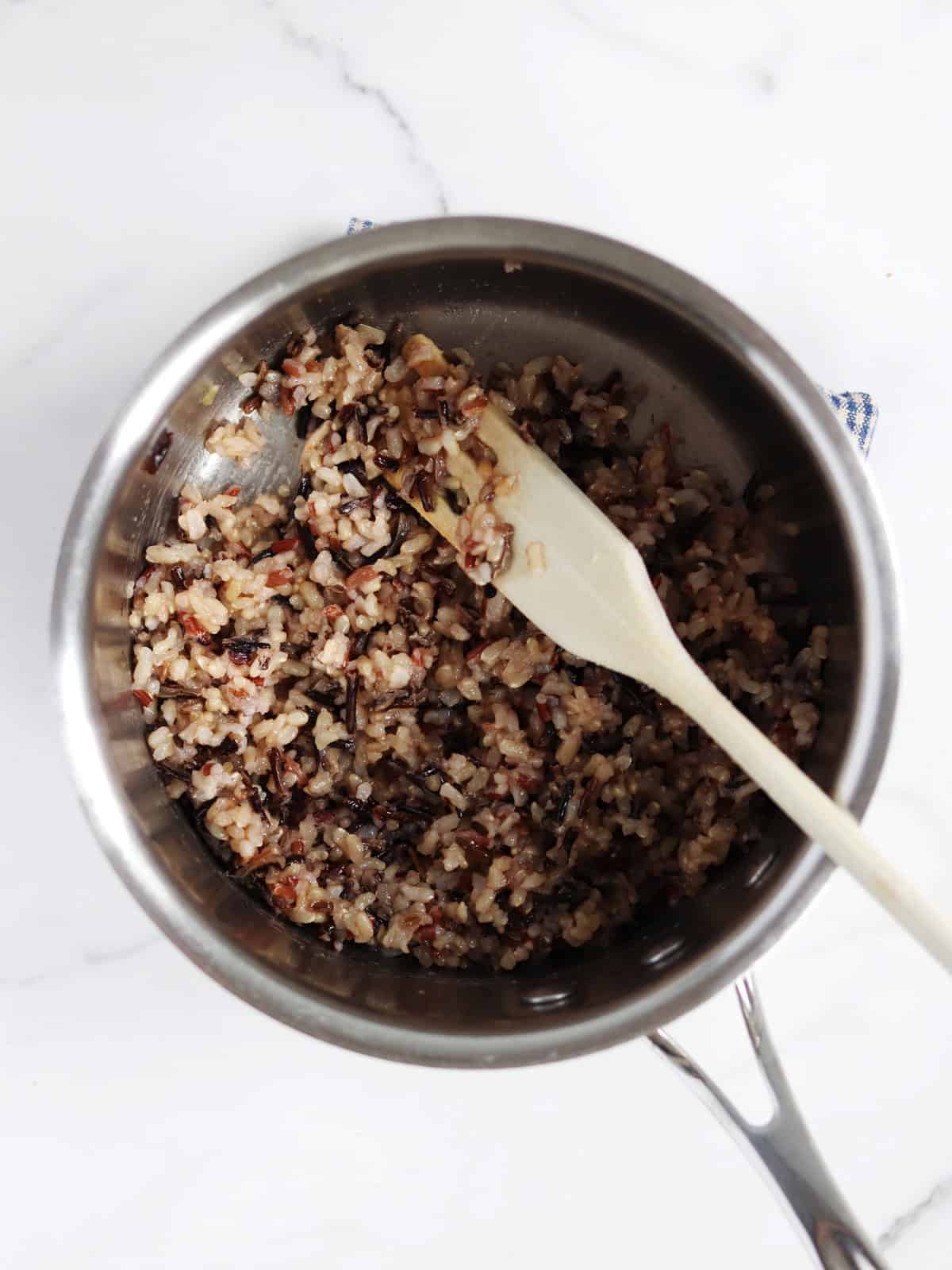 A stainless steel pot filled with cooked wild rice, being stirred with a wooden spoon, sits on a white marble countertop.