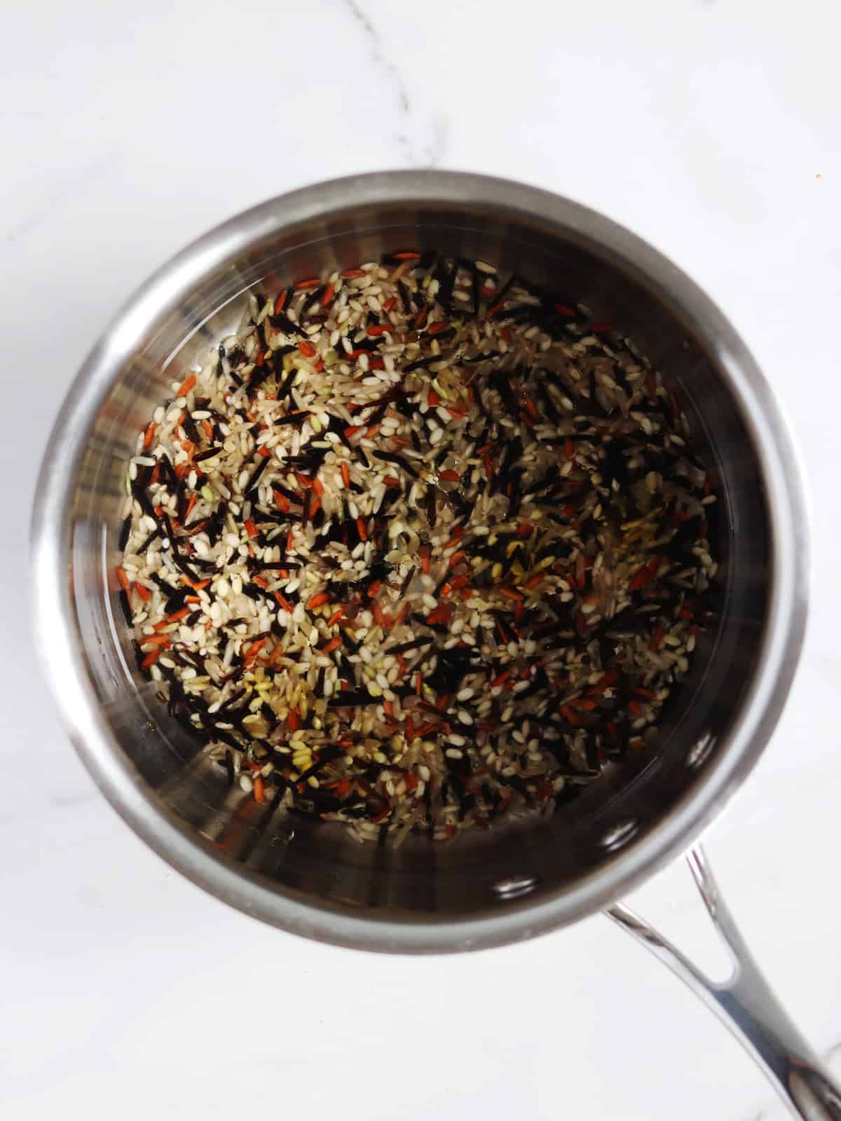 A stainless steel pot containing a mix of uncooked wild rice sits on a white marble surface.