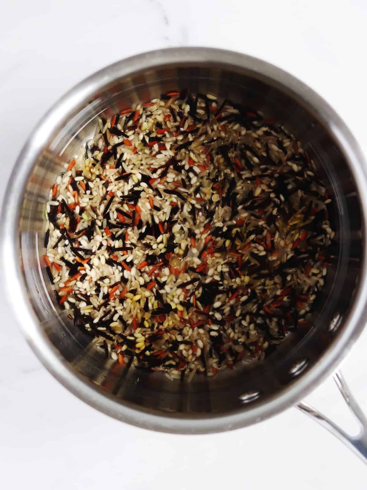 A stainless steel pot filled with uncooked wild rice blend, showing a mix of brown, black, and red rice on a white marble countertop.