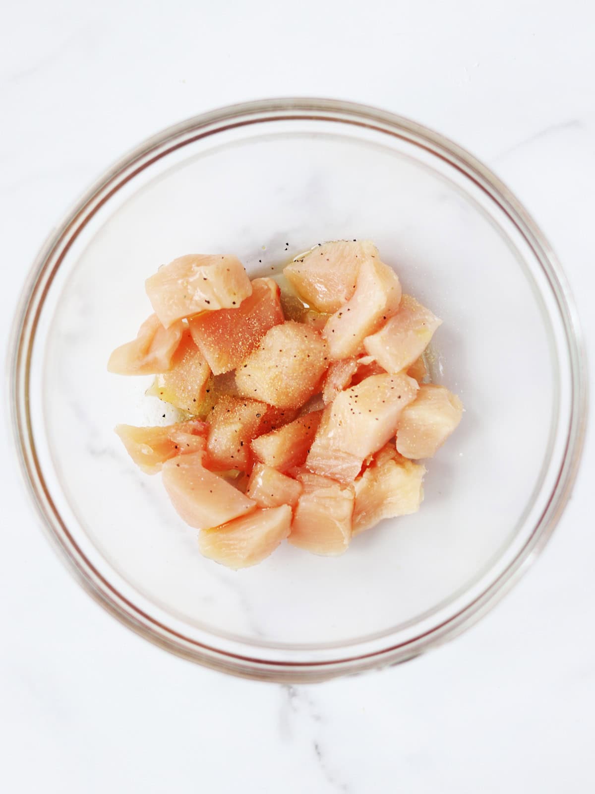 Raw, cubed chicken pieces seasoned with garlic powder, salt and pepper in a clear glass bowl on a white marble surface.