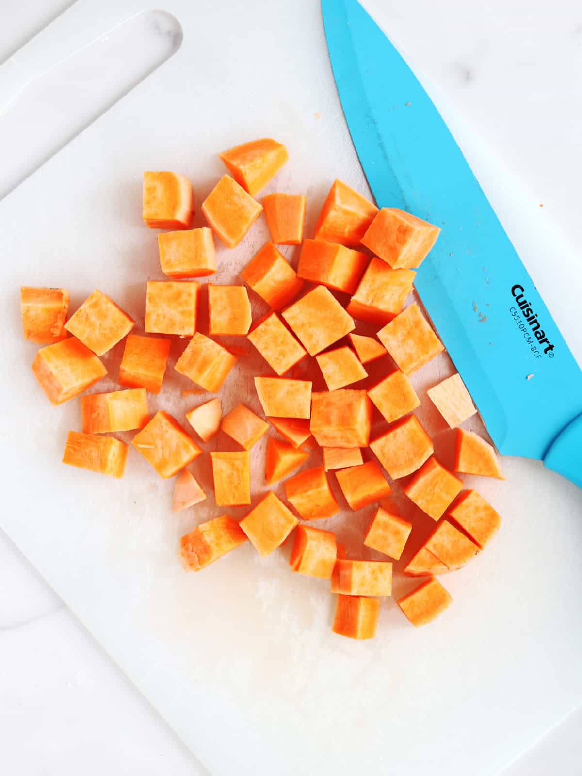 Diced sweet potato pieces on a white cutting board next to a blue-handled kitchen knife.