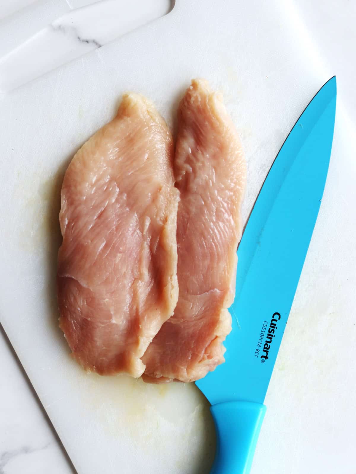 Two raw chicken breast halves placed side by side on a white cutting board next to a ceramic blue chef's knife.