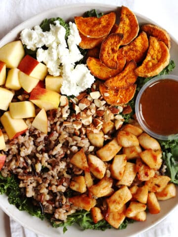 A Harvest Bowl with diced apples, crumbled goat cheese, roasted sweet potato slices, chopped almonds, wild rice, seasoned chicken pieces, kale, and a small container of balsamic vinaigrette dressing.