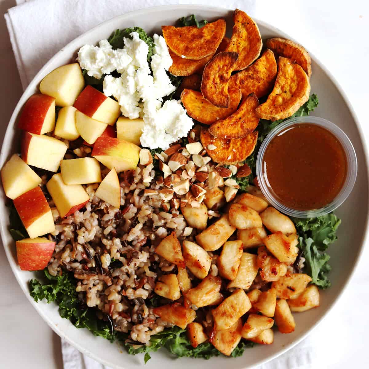 A Harvest Bowl with diced apples, crumbled goat cheese, roasted sweet potato slices, chopped almonds, wild rice, seasoned chicken pieces, kale, and a small container of balsamic vinaigrette dressing.
