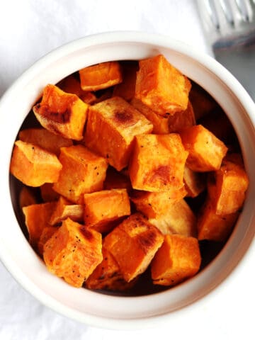 A white bowl filled with roasted sweet potato cubes sits on a white napkin next to a silver fork.