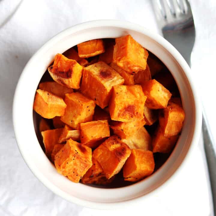 A white bowl filled with roasted sweet potato cubes sits on a white napkin next to a silver fork.