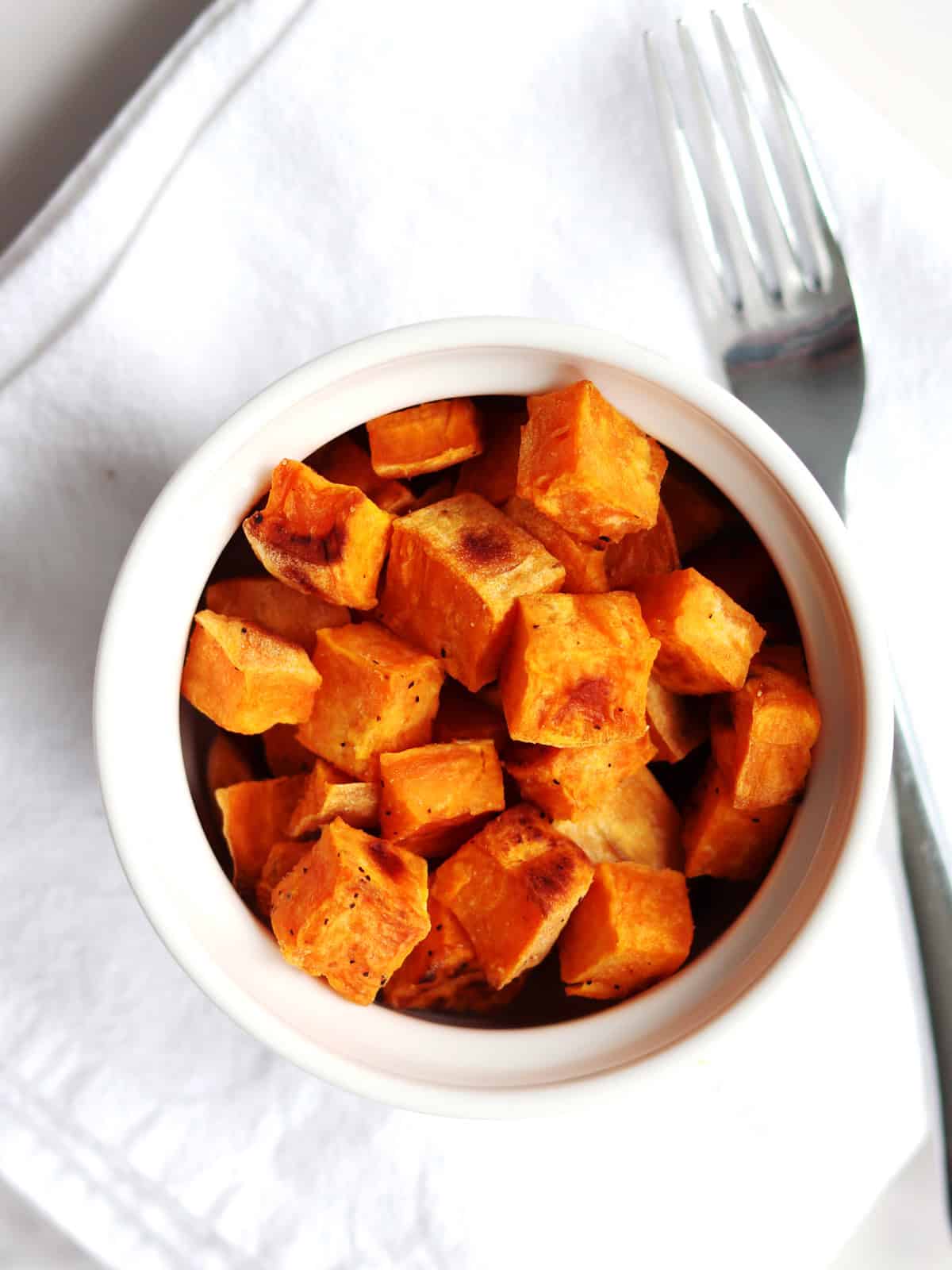 A white bowl filled with roasted sweet potato cubes sits on a white napkin next to a silver fork.