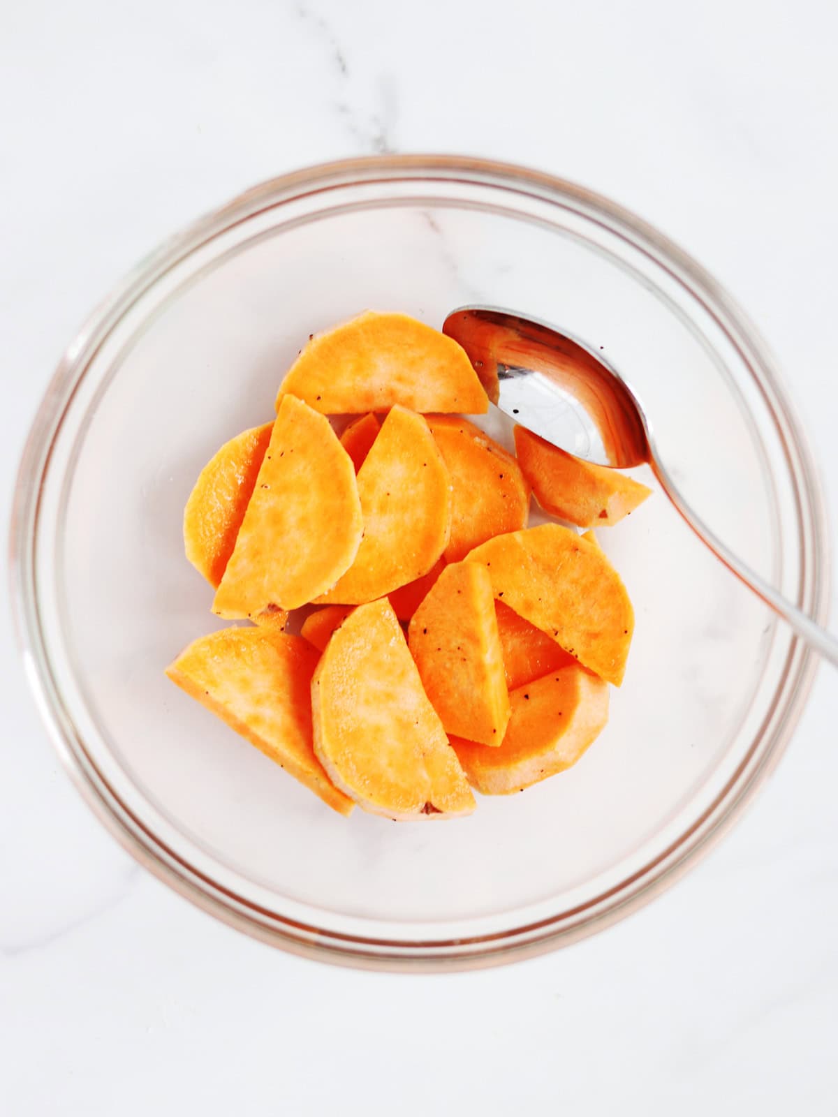A glass bowl containing sliced sweet potatoes, with a metal spoon resting inside, on a white marble surface.