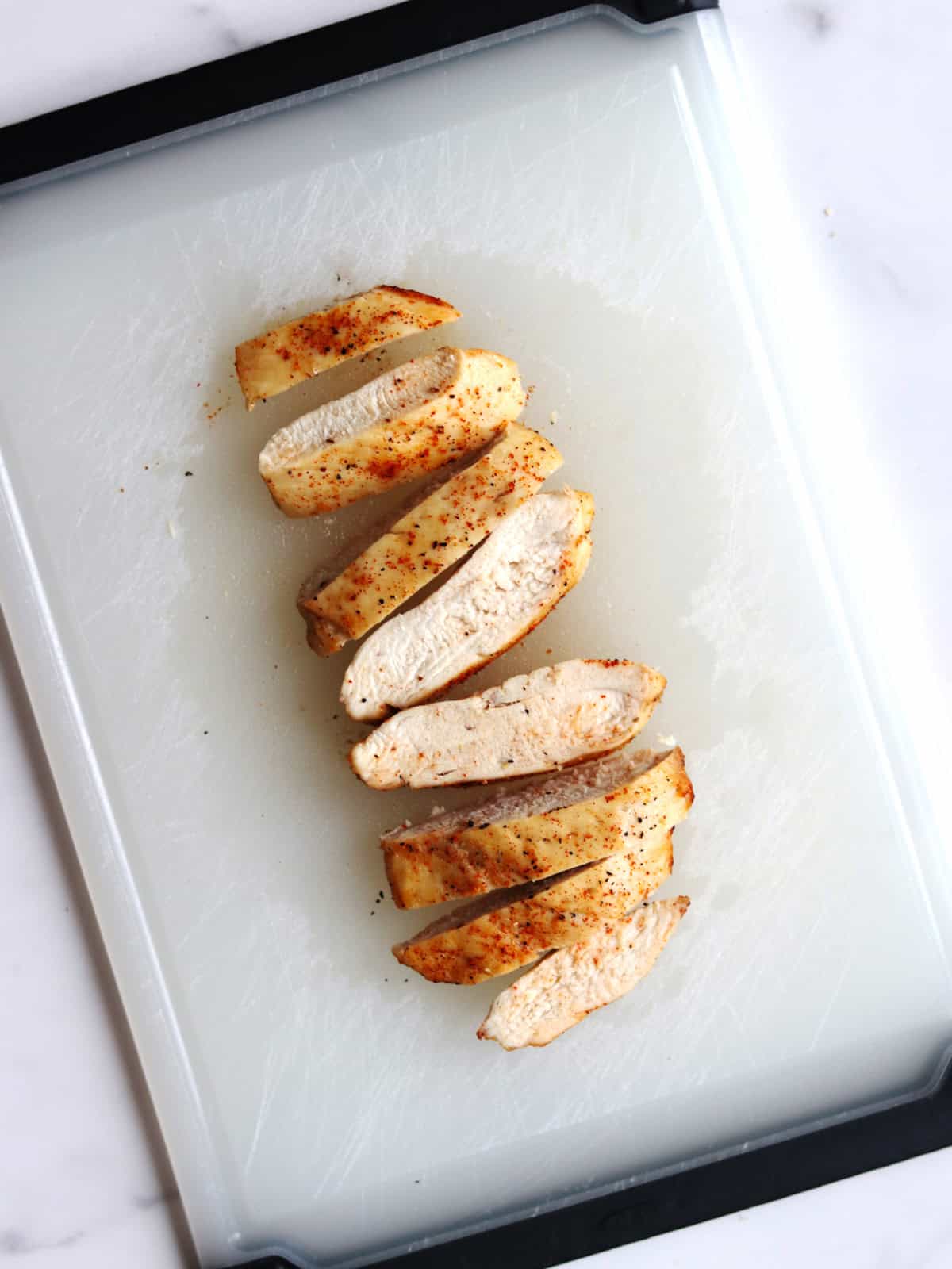 Sliced cooked air fryer chicken breast, seasoned with spices, arranged neatly on a white cutting board with a black border on a light surface.