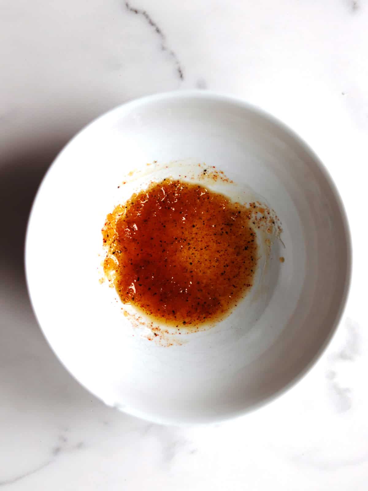 A small amount of sweet and savory seasoning rub with visible spices sits at the bottom of a white bowl on a marble countertop.