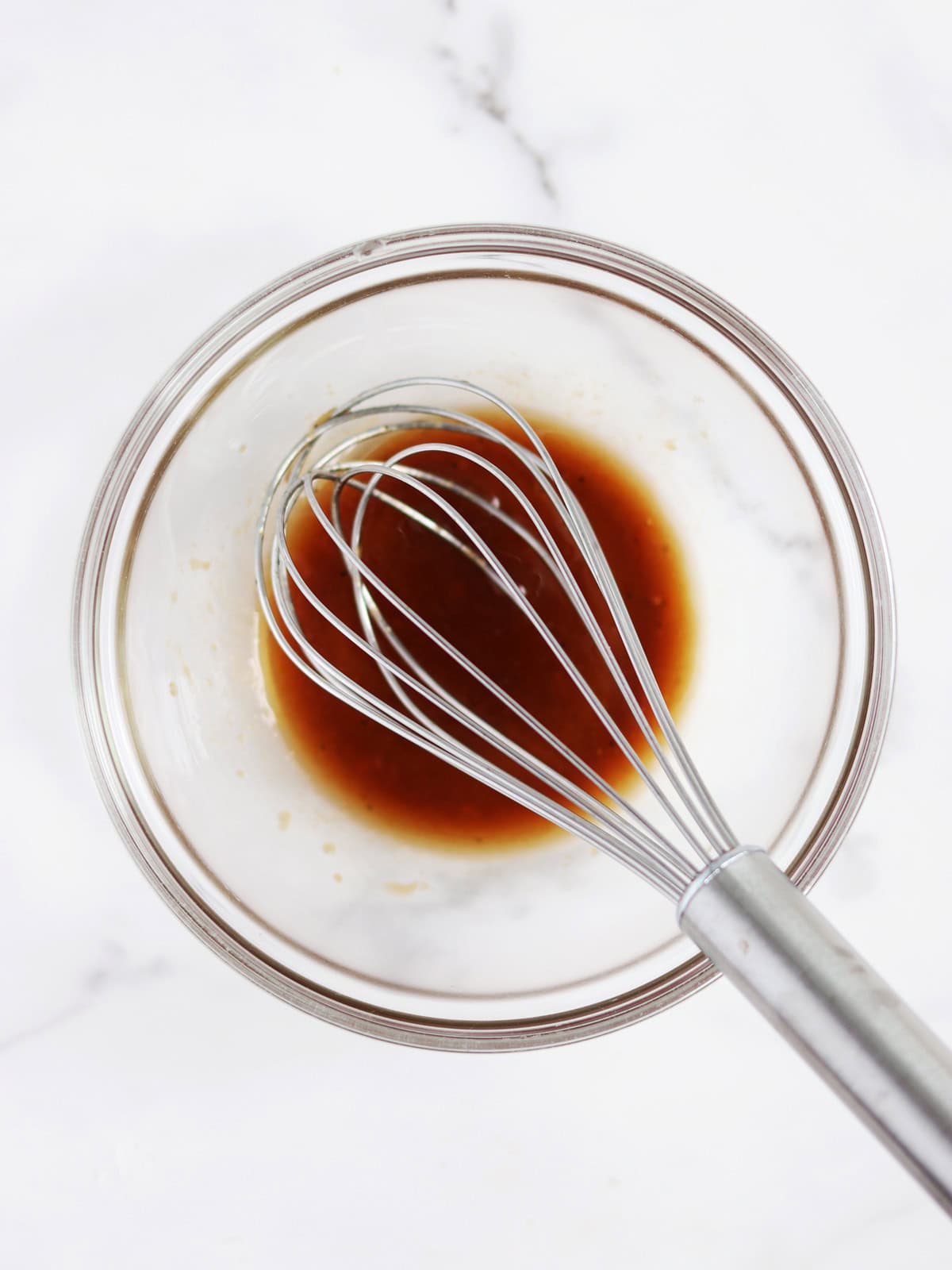 A metal whisk rests in a glass bowl containing a small batch balsamic vinaigrette on a white marble countertop.