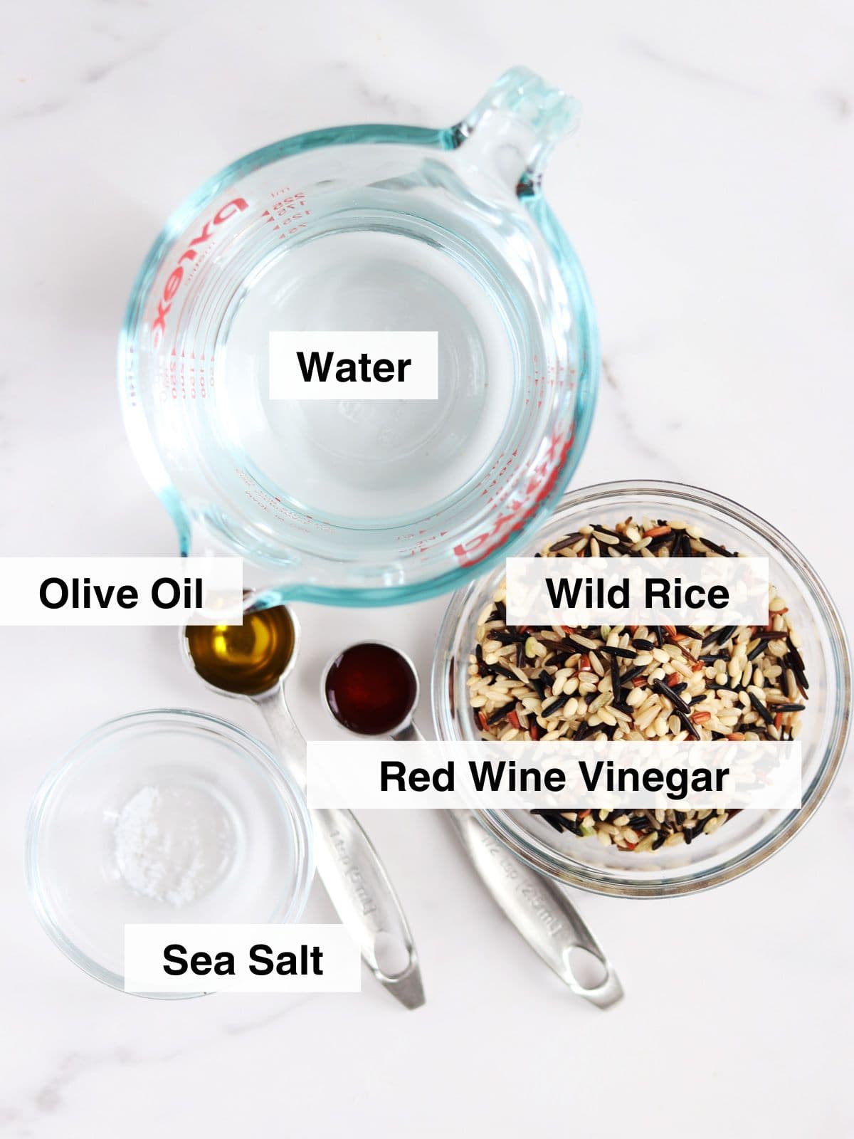 Ingredients for wild rice including a glass measuring cup of water, a bowl of wild rice, a spoon of olive oil, a spoon of red wine vinegar, and a spoon of sea salt are arranged on a white marble countertop, each labeled with text.