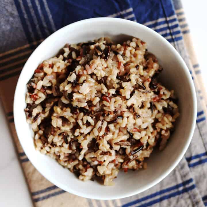 A white bowl filled with cooked wild rice sits on a tan napkin with a dark blue plaid pattern.