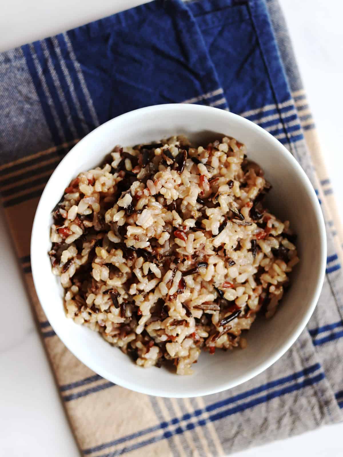 A white bowl filled with cooked wild rice sits on a blue and tan plaid napkin on a white marble countertop.