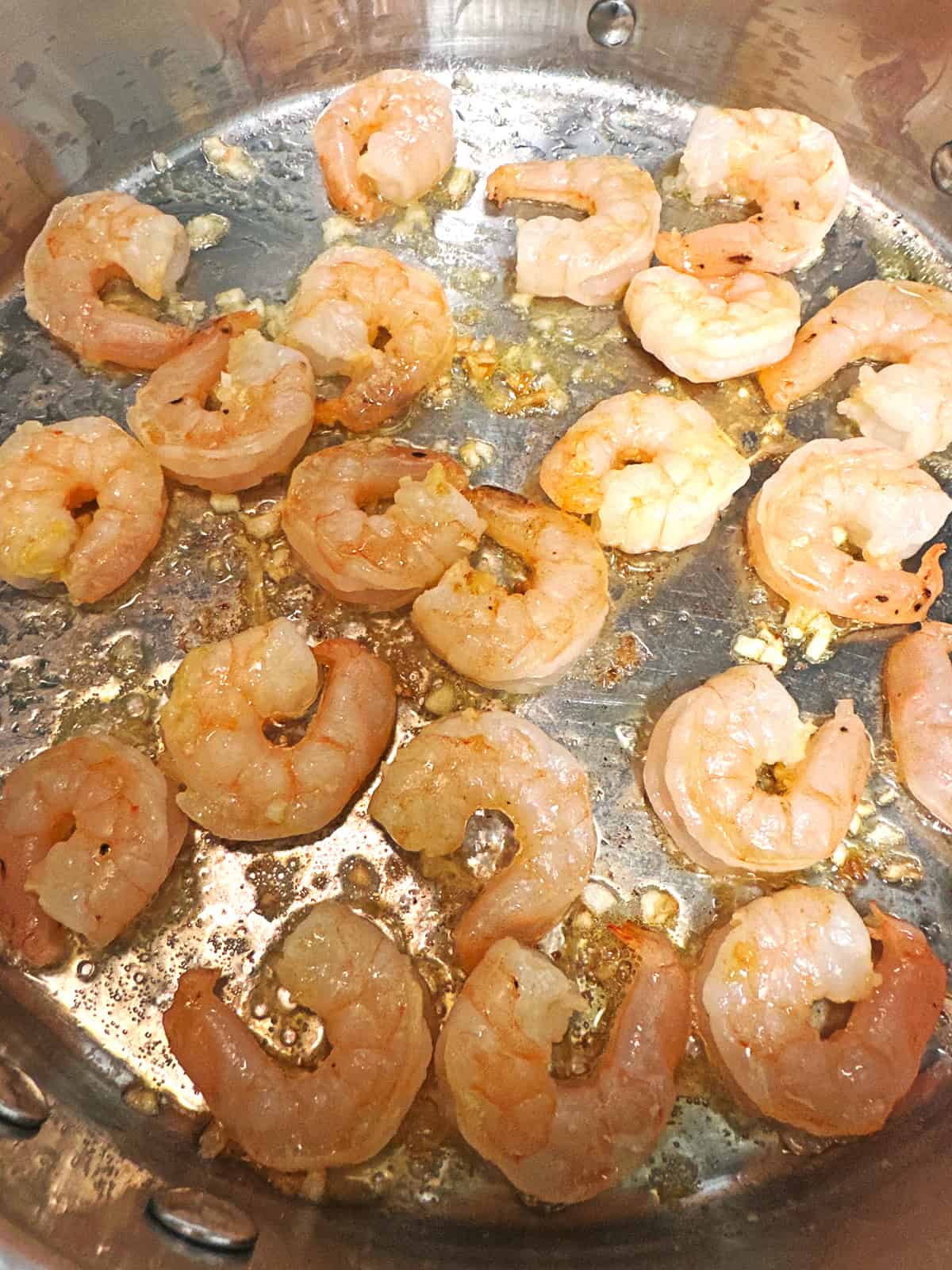 Shrimp cooking in a stainless steel pan with bits of minced garlic and oil, showing a slightly pink color as they sauté.